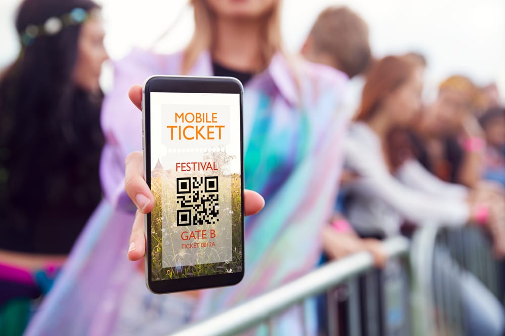 Person holding smartphone with mobile ticket QR code for festival entry, amid lively crowd in vibrant event setting.