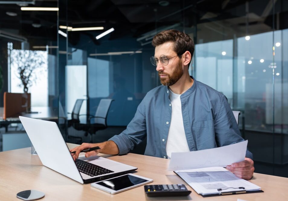 Focused IT manager in modern office analyzing data on laptop, holding document at desk with smartphone and calculator.