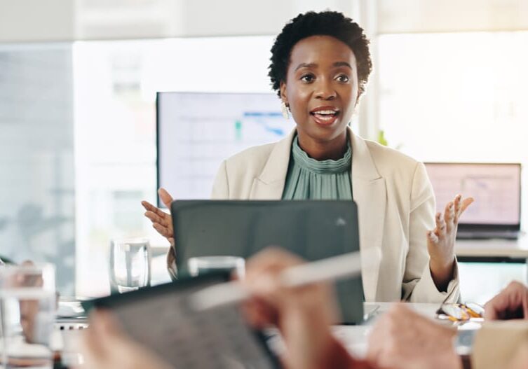 Woman in blazer gesturing during risk assessment meeting at table with laptops, colleagues listening in modern office.